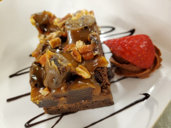 A turtle brownie plated with caramel and toasted pecans, a strawberry and a chocolate drizzle alongside.