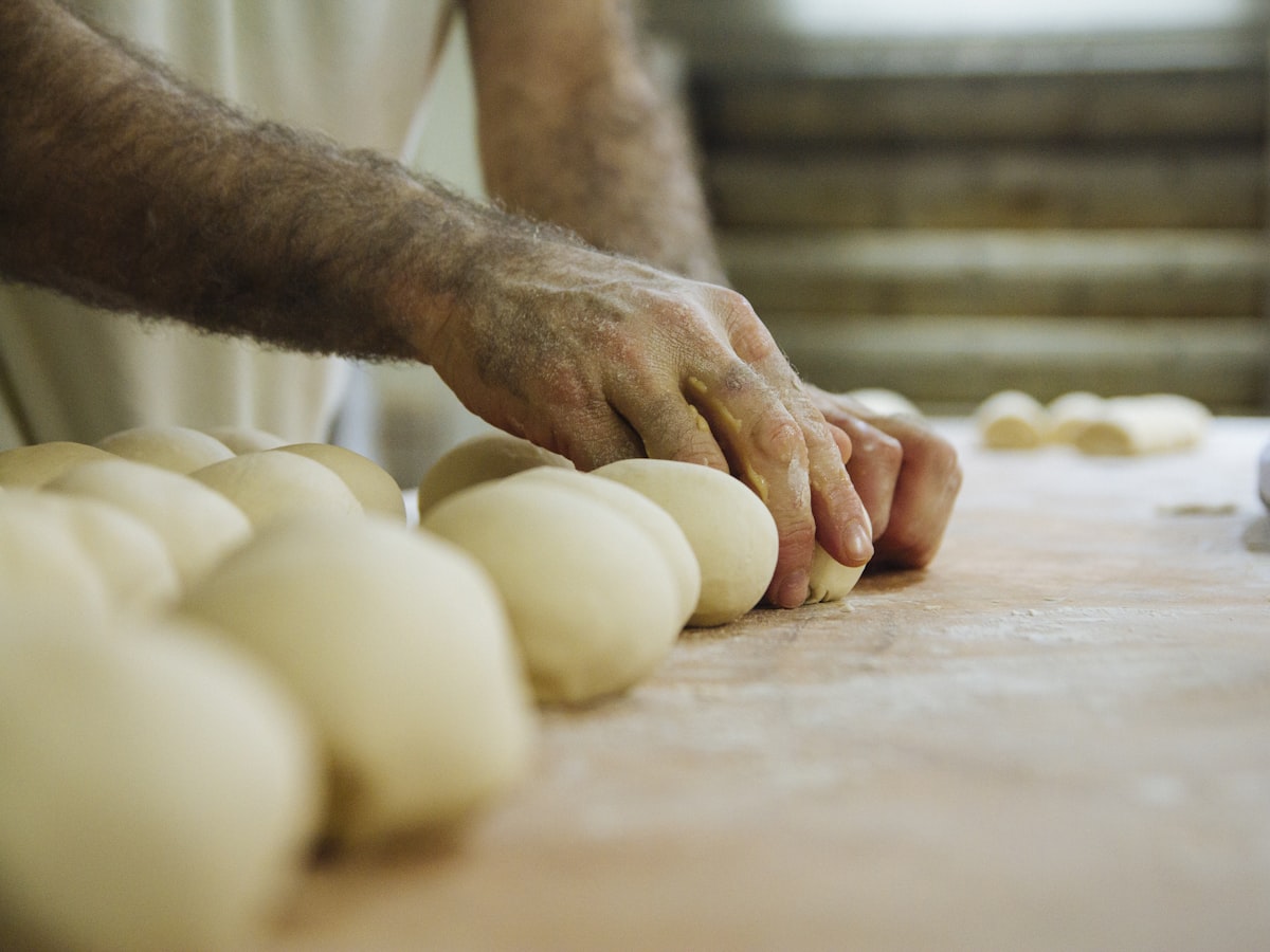 Floured hands rolling a row of dough rounds along a wooden bench.
