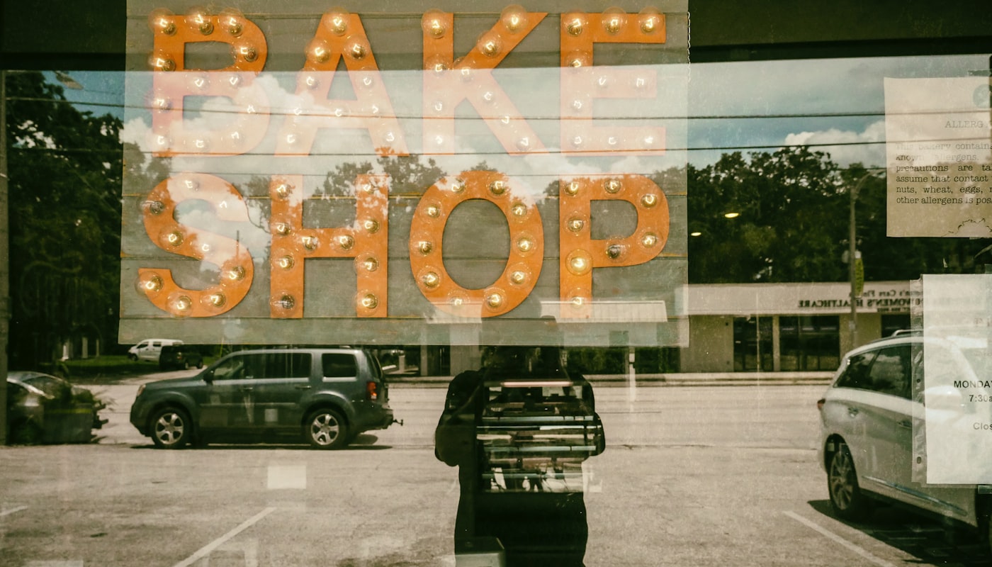 A lit-bulb marquee sign reading BAKE SHOP, seen through the front window of the bakery.