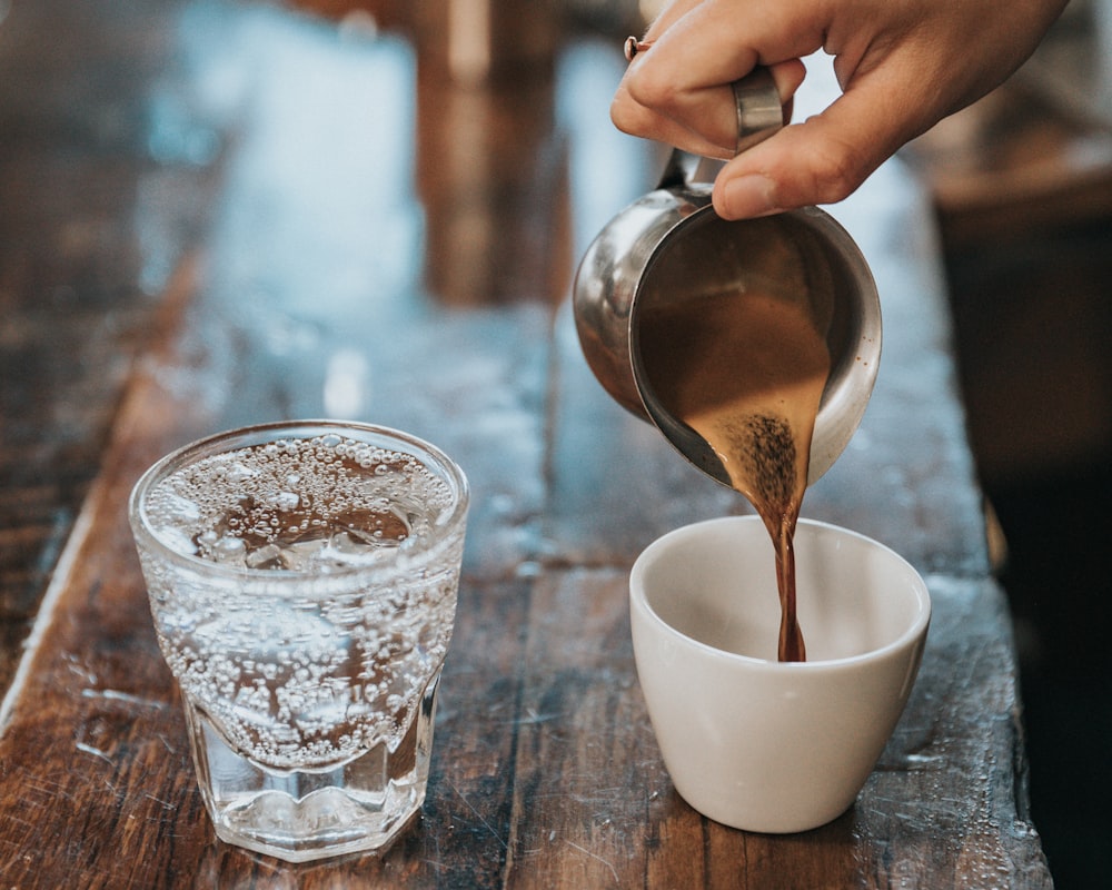 A fresh espresso shot being poured from a steel pitcher into a small white cup, sparkling water alongside.
